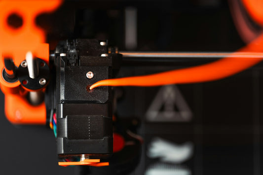 Close-up of a 3D printer nozzle extruding orange filament, illustrating indoor 3D printing safety